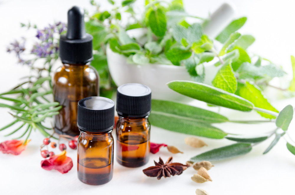 3 small essential oil bottles next to a white mortar and pestle surrounded by green leaves, herbs, purple flowers, and red berries. 