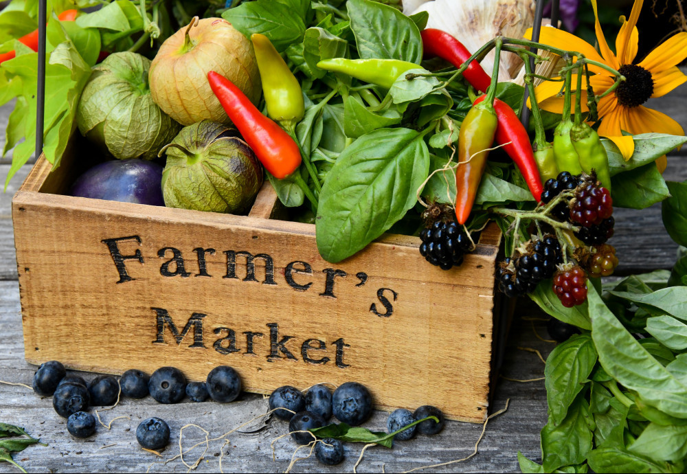 A wooden box that reads Farmer's Market filled with fresh vegetables and a yellow flower and fresh blueberries next to it. 