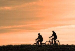 The dark silhouettes of 2 people riding bikes in front of an orange sunset. 