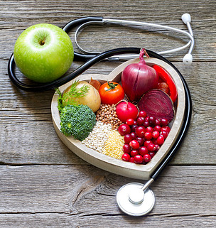 A wooden bowl in the shape of a heart filled with vegetables and seeds surrounded by a stethoscope with a green apple next to it. 