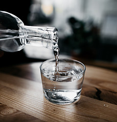 A small glass of water with a glass bottle of water being poured into it on a wooden counter. 