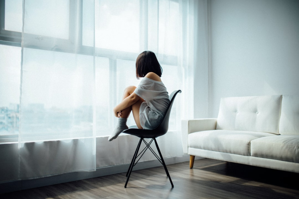 A woman in pajamas sitting in a chair looking out of the window longingly holding her knees at her chest. 