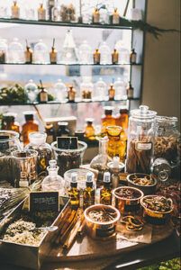 Many glass bottles, jars, and boxes filled with herbs, tinctures, and natural medicines on a table in front of a window. 