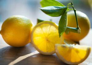 2 whole lemons and 2 lemon slices on a wooden counter with green leaves next to them. 