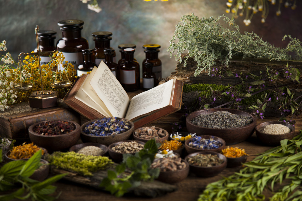An opened book amongst bowls of herbs and dried flowers next to several bottles of tinctures of natural medicines and herbs. 