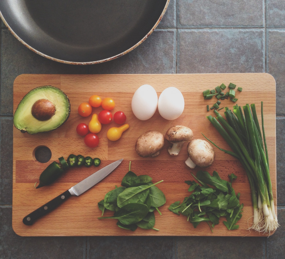 A wood cutting board with fresh vegetables and 2 whole eggs on it next to an empty pan on a tile counter. 