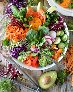 A vibrantly colored bowl of salad with fresh avocado, carrots, and cabbage surrounding it on a wooden surface. 