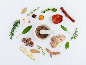 A mortar and pestle with brown seeds in it surrounded by green herbs, ginger root, and a cinnamon stick. 