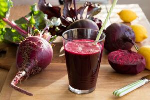 A glass of bright pink beet juice with a straw surrounded by fresh beets and lemons, some whole, some sliced on a wooden cutting board.