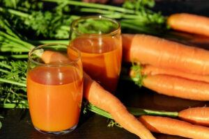 Two small glasses of bright orange carrot juice surrounded by large whole carrots with green tops. 