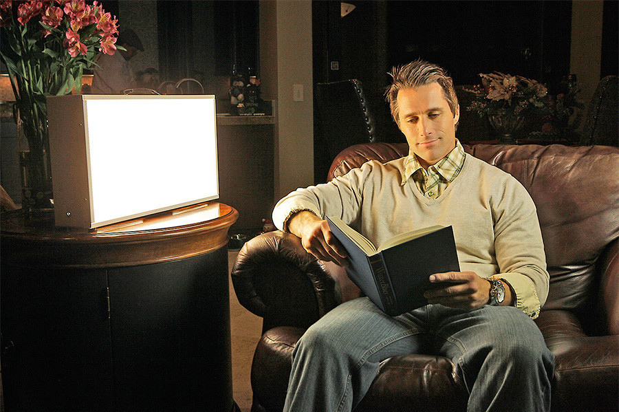 A man sitting in front of a large light therapy box that is emiting bright light reading a book. 