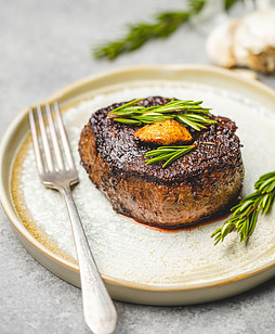 A filet mignon steak on a tan circular plate with green rosemary stalks as a garnish on top and a silver fork resting on the plate next to the steak.