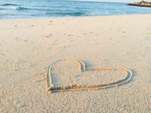 A beach scene with a heart drawn in the sand in the foreground. 