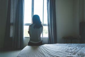 The back of a woman sitting on a bed facing a window with her arms crossed.