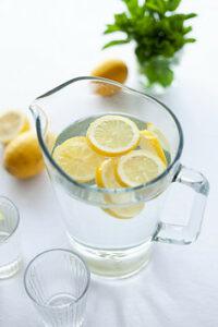 A glass pitcher of water with slices of lemon in it surrounded by 2 glasses, lemons, and green leaves.