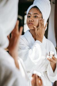 A woman looking in the mirror with a robe on and towel on her head applying lotion to her face.