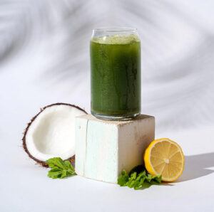 A glass of green juice with coconut and lemon next to it. 