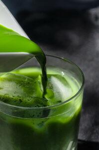 A glass of green juice with more green juice being poured into it.