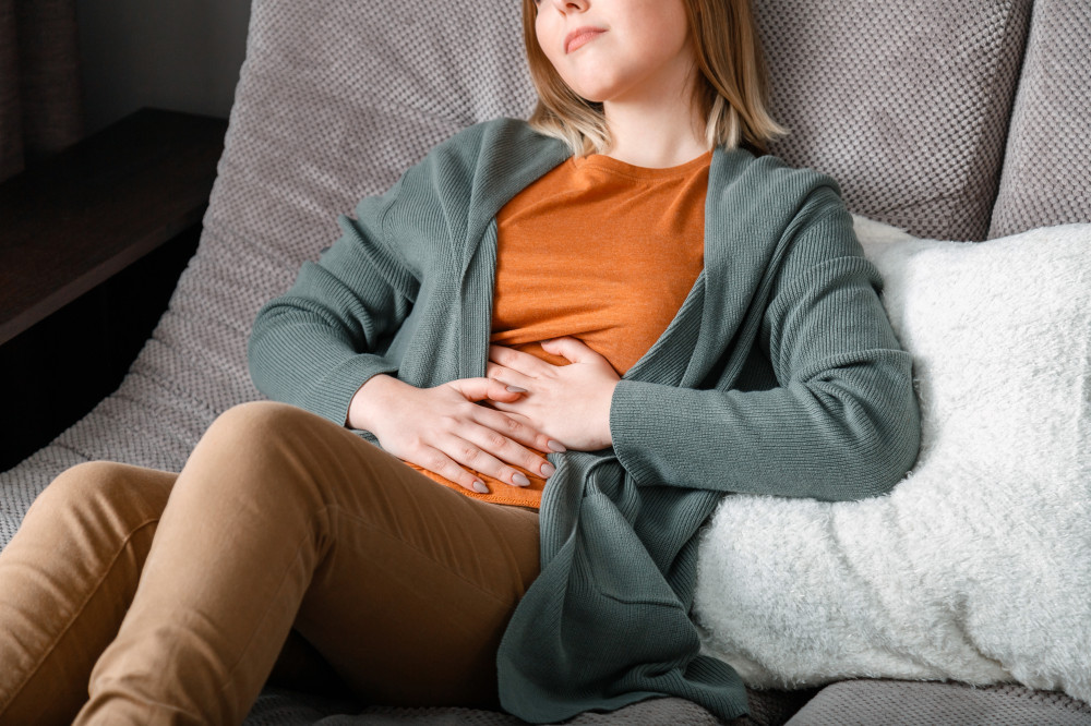 A woman sitting on a couch with her hands on her abdomen experiencing bloating.